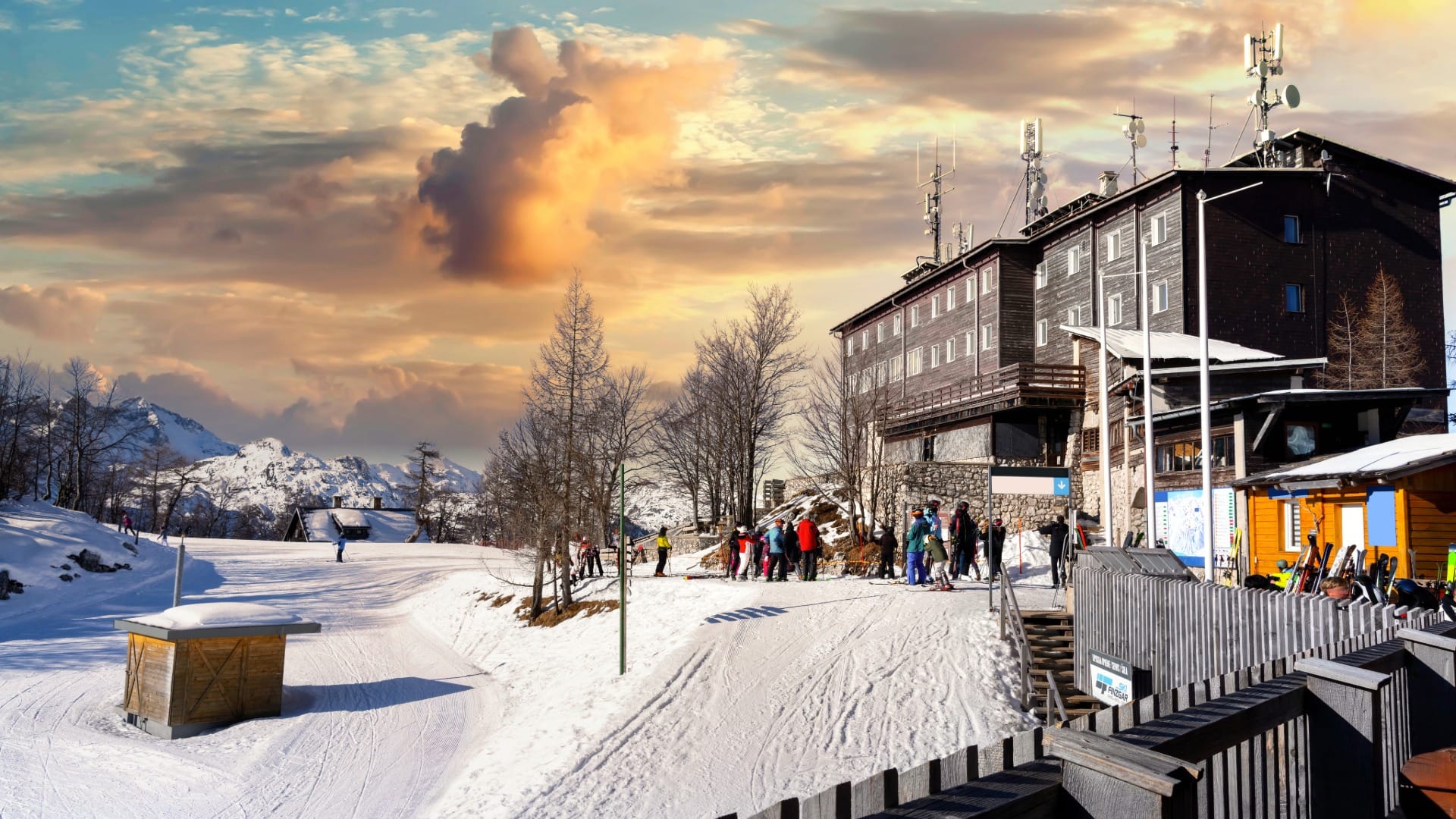 Wooden ski lodge terrace crowded with skiers below snowy mountains