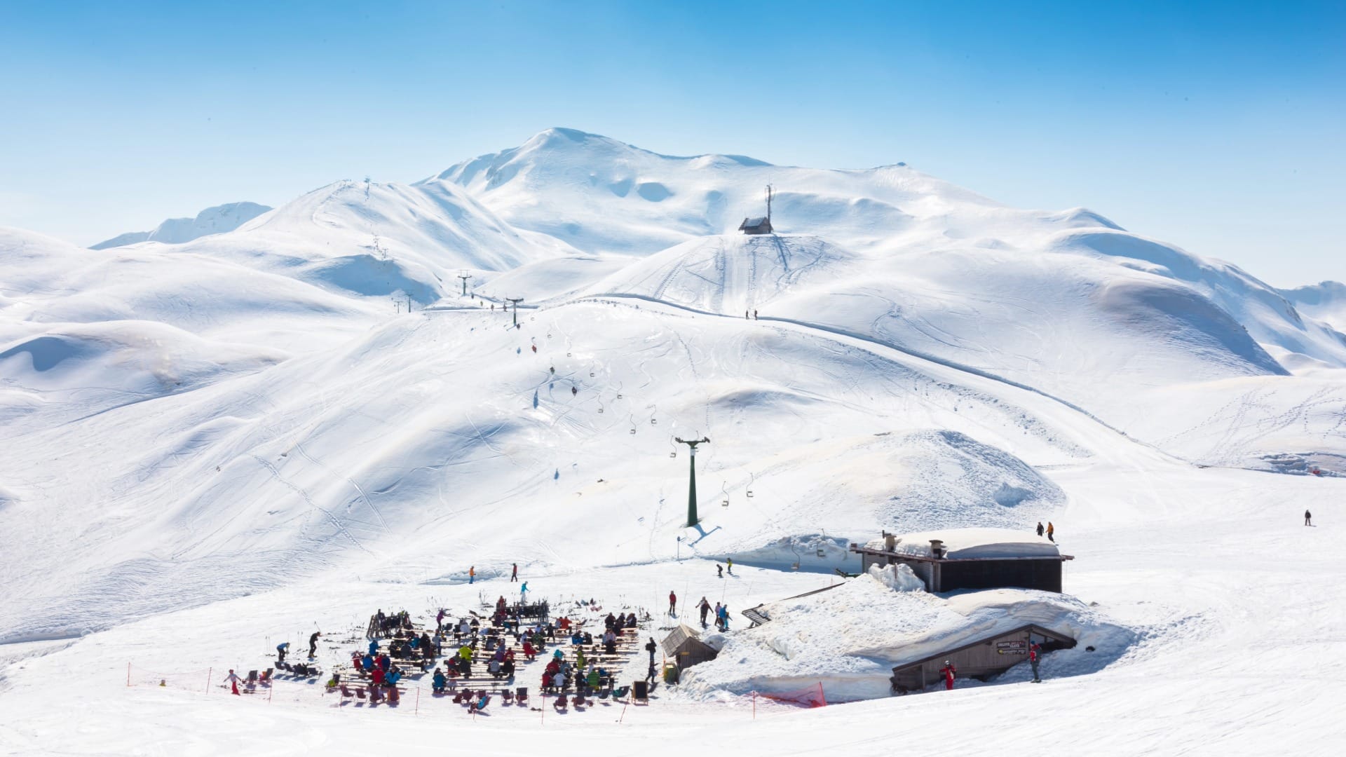Snow-covered mountain-top terrace crowded with people at picnic tables between runs
