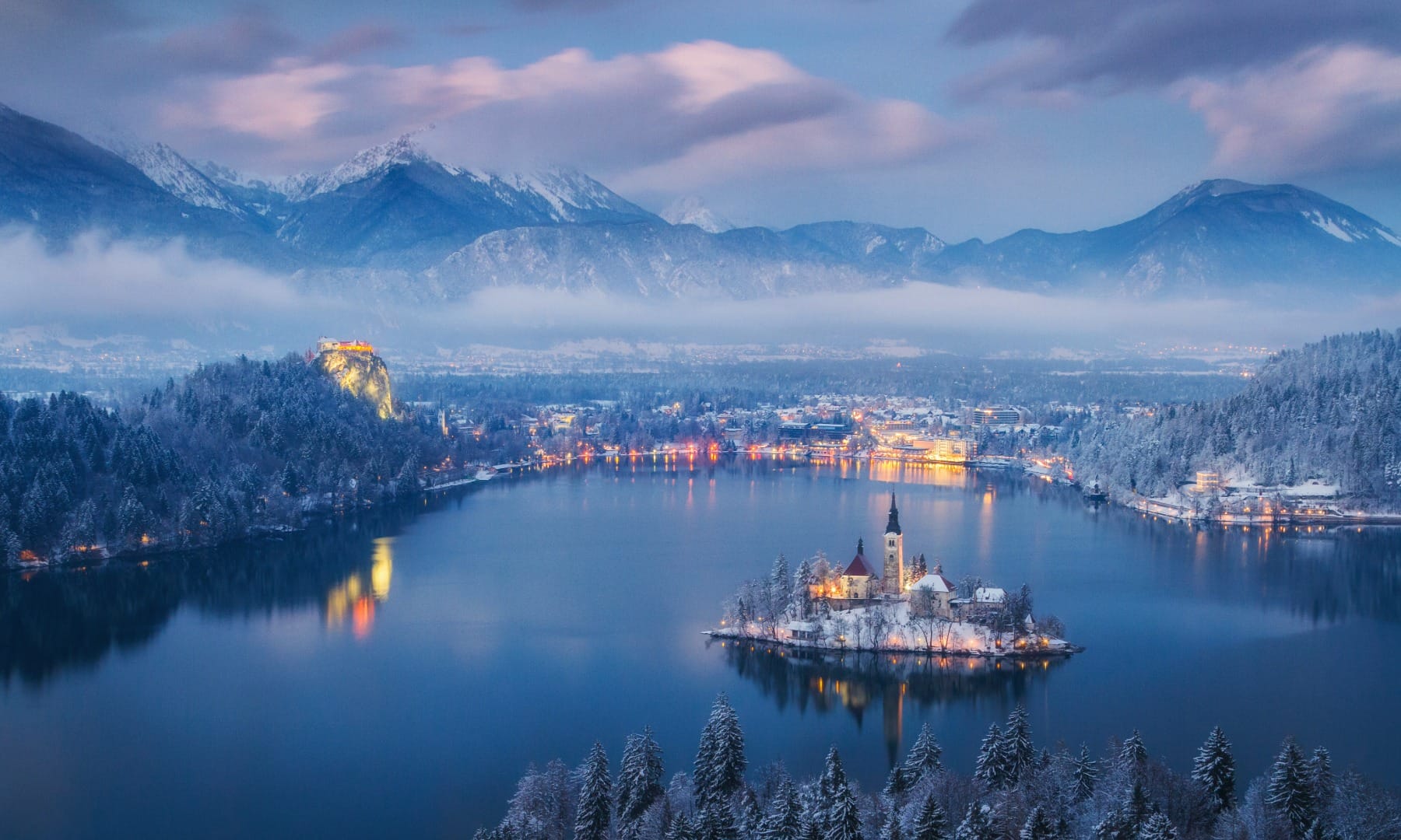 Aerial dusk view of Lake Bled with the island church lit up, the castle on its cliff and snowy mountains behind