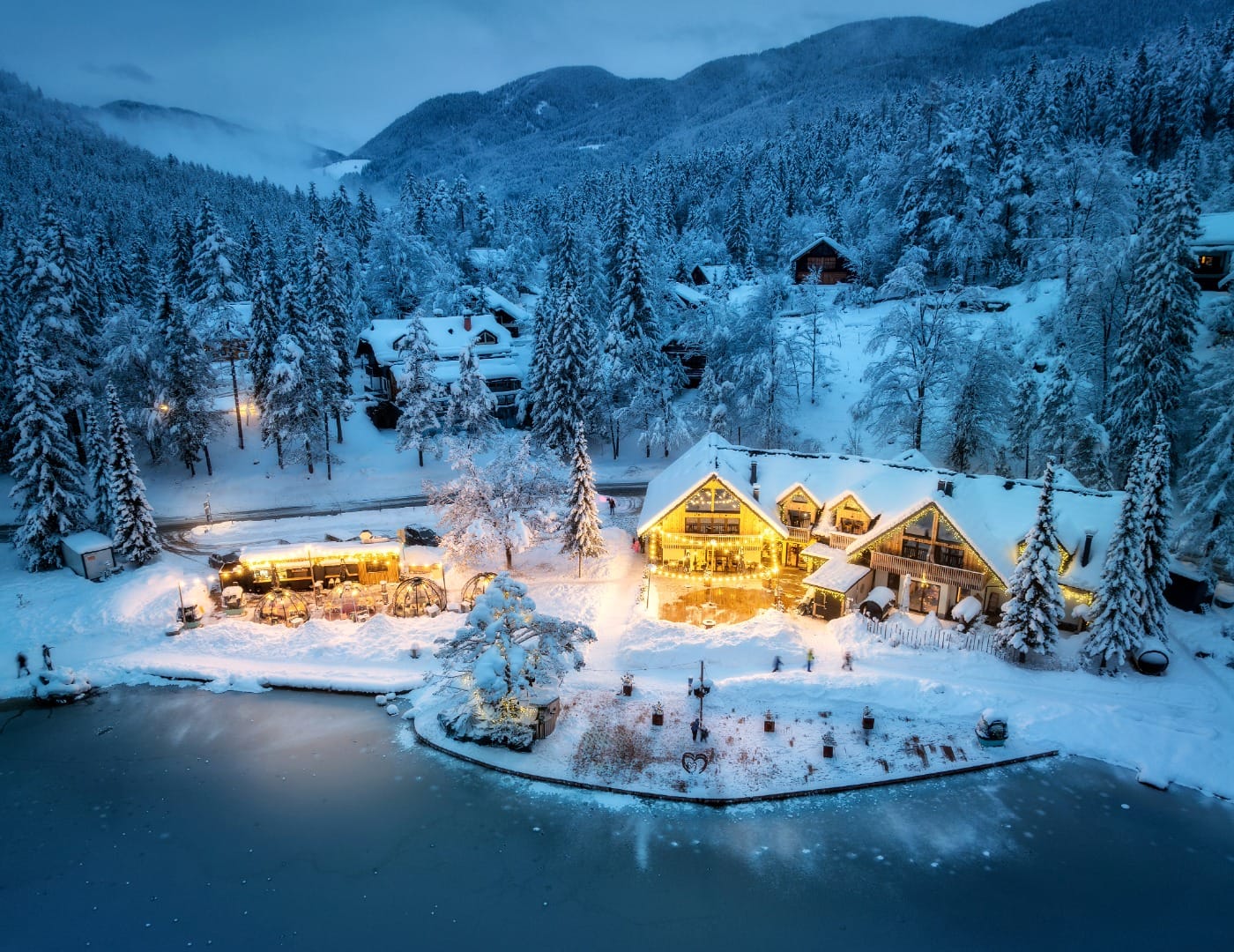 Snowy alpine lodge and cabins at dusk beside a partially frozen lake with cosy lit windows