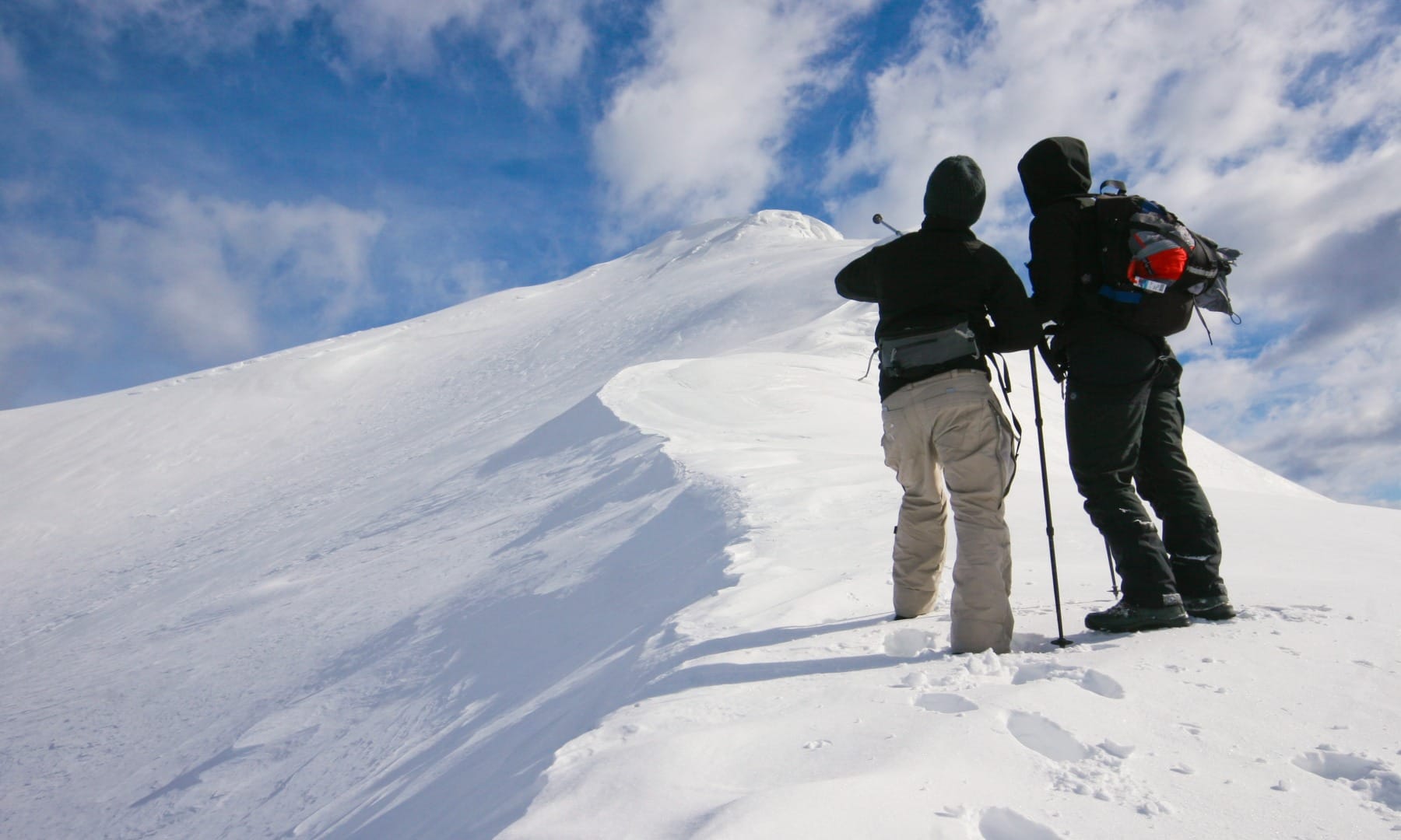 Two hikers pausing near a snowy mountain summit ridge in the Julian Alps