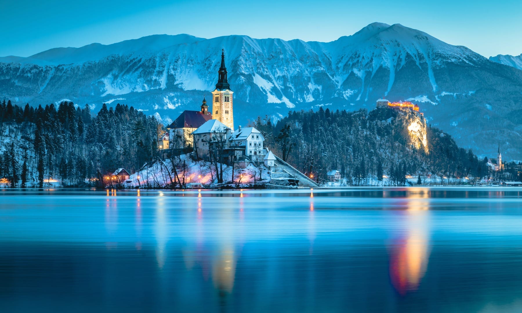 Bled Island church lit up at winter dusk with Bled Castle on the cliff behind and reflections on the lake