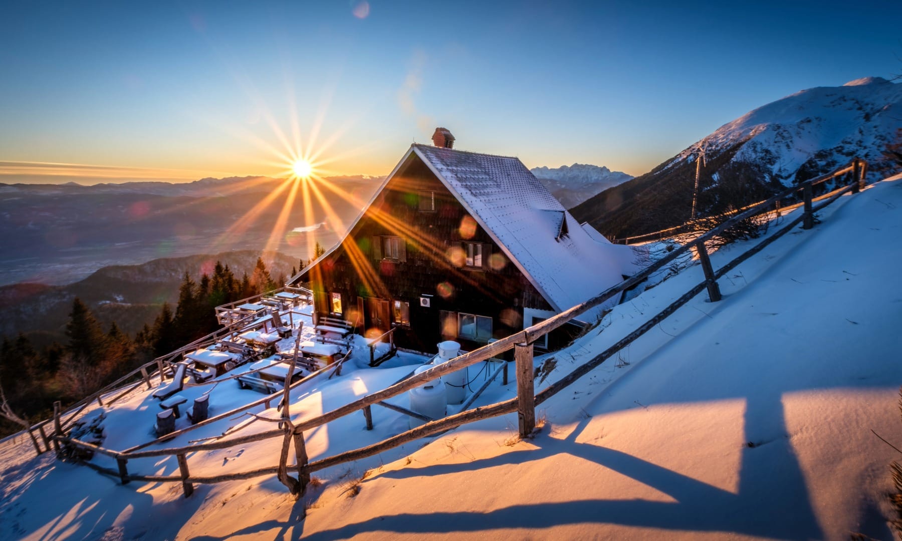 Snow-covered mountain hut terrace at sunrise with sun flare over the valley