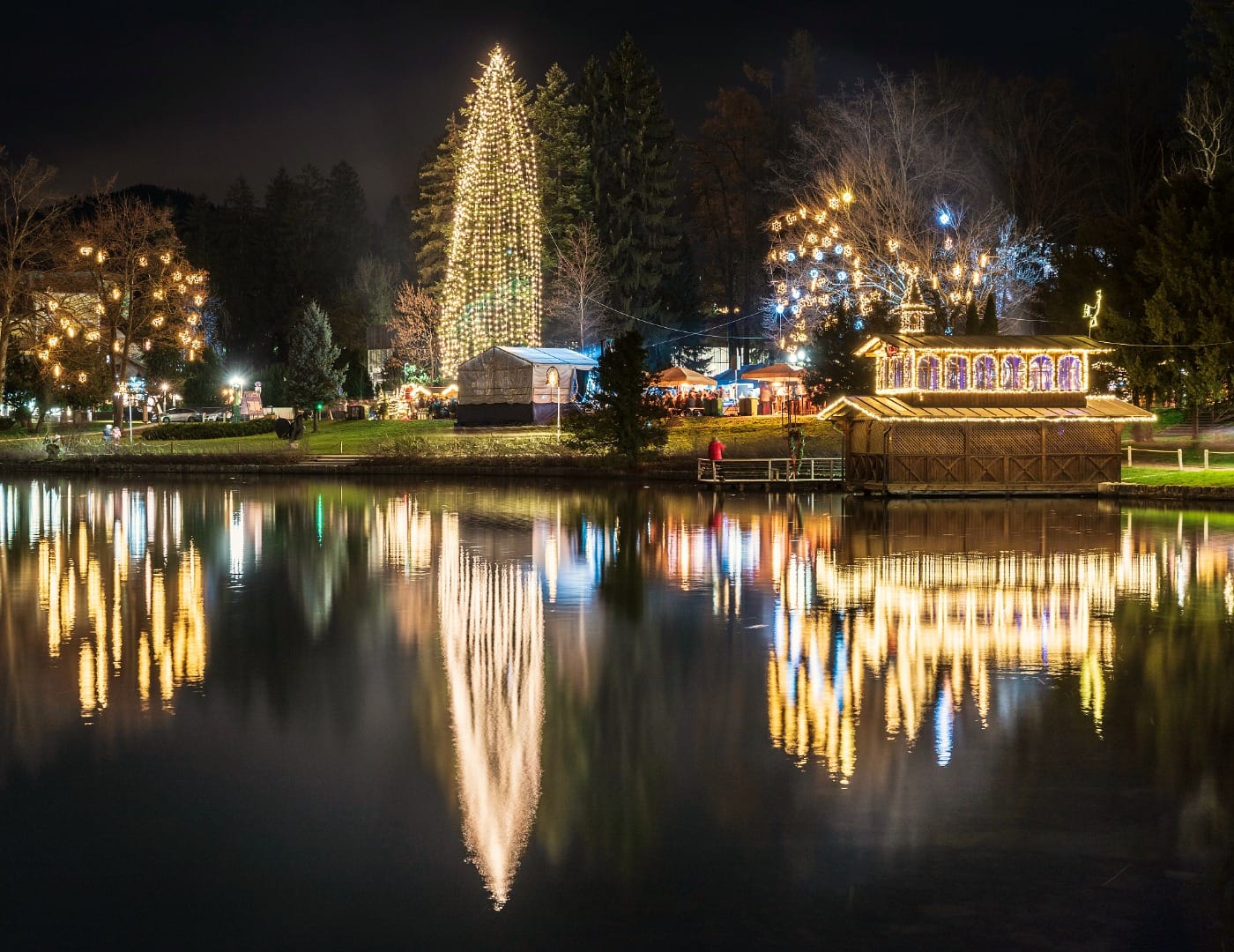 Tall lit Christmas tree reflected on Bled lake at night with market stalls nearby