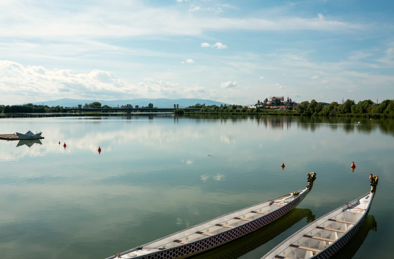 Two dragon boats moored on the still water of Ptuj Lake, with Ptuj Castle on its hill in the distance