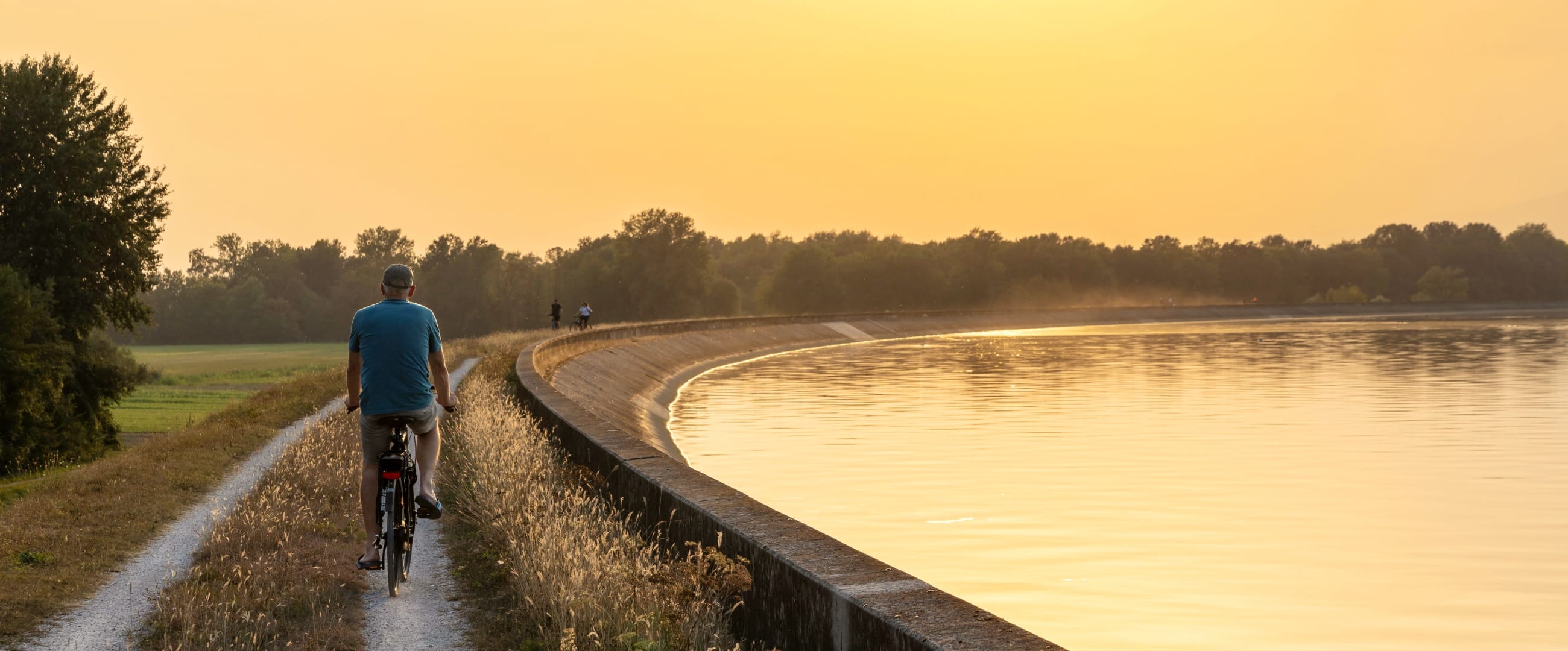 A cyclist on a gravel path along the curved concrete flood embankment of a wide reservoir at sunset