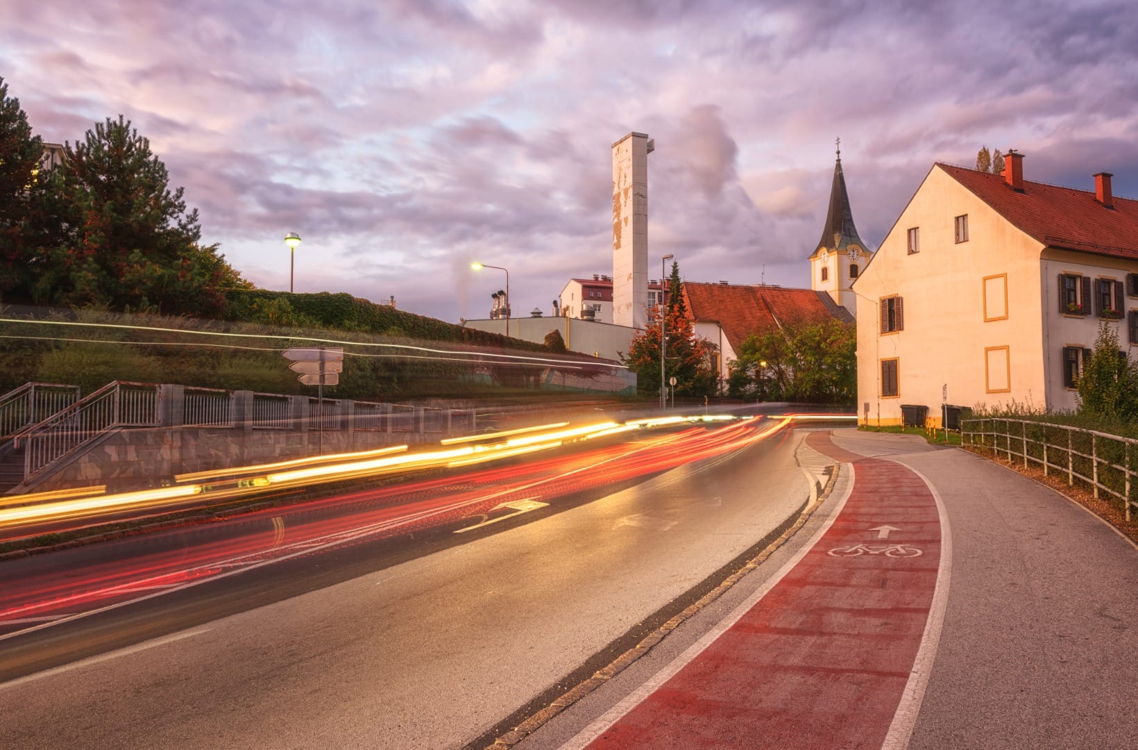 A red-painted on-road cycle lane running past a village church at dusk, car light trails on the adjacent road