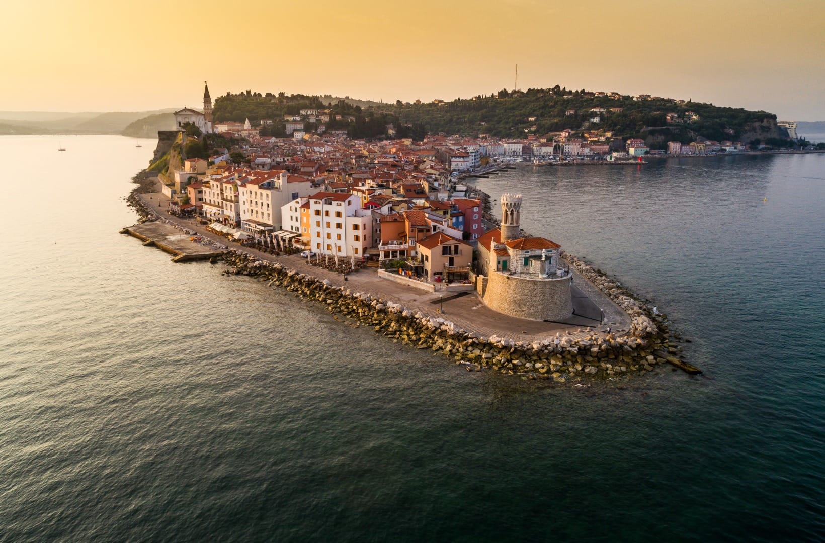 Aerial view of Piran at sunset, red rooftops packed onto a narrow peninsula with the church of St George above