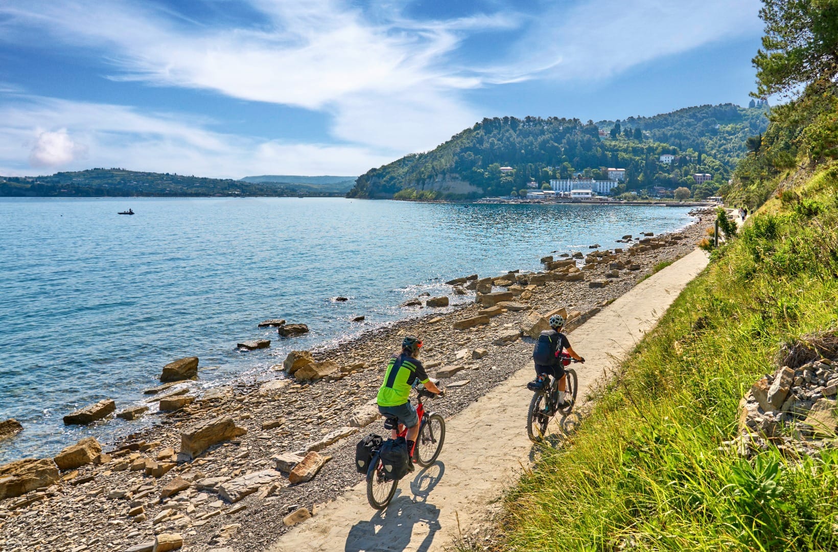 Two cyclists with panniers riding a gravel path along a rocky shoreline, the Strunjan peninsula ahead