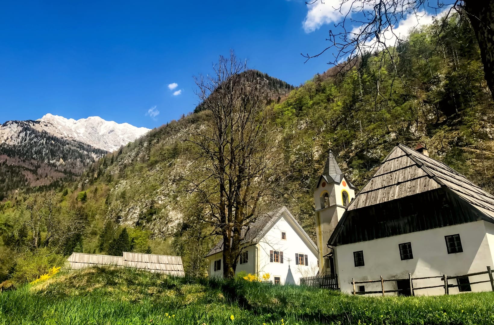 A small alpine church and an old farmhouse with a wooden roof below a snow-streaked peak in the Soča valley