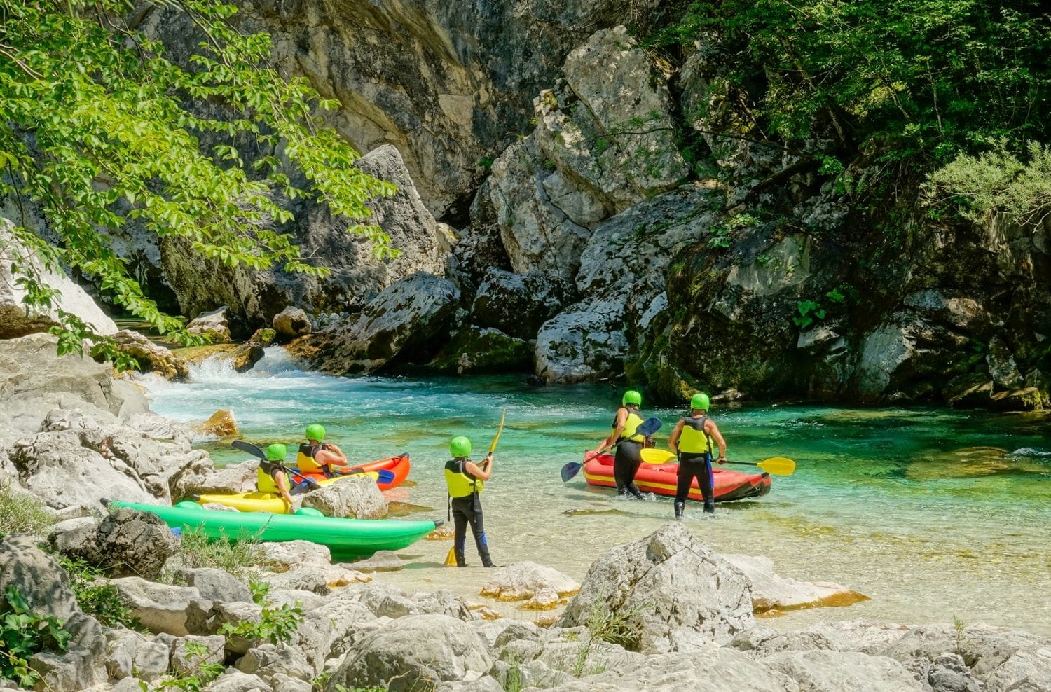 Small group with inflatable kayaks on a white gravel beach beside a low cascade in a rocky bend of the Soča