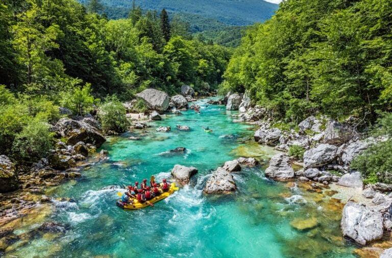 Yellow raft with a paddling crew running a whitewater rapid on the turquoise Soča between forested banks, seen from above
