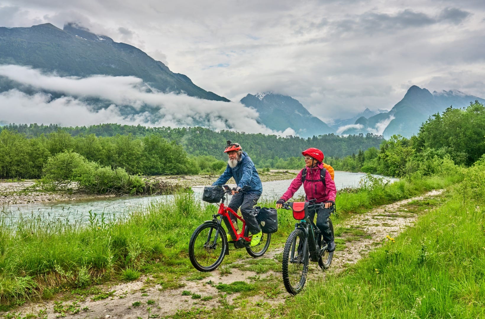 Two cyclists with panniers on a gravel path beside the Soča river, cloud-draped peaks of the Julian Alps behind
