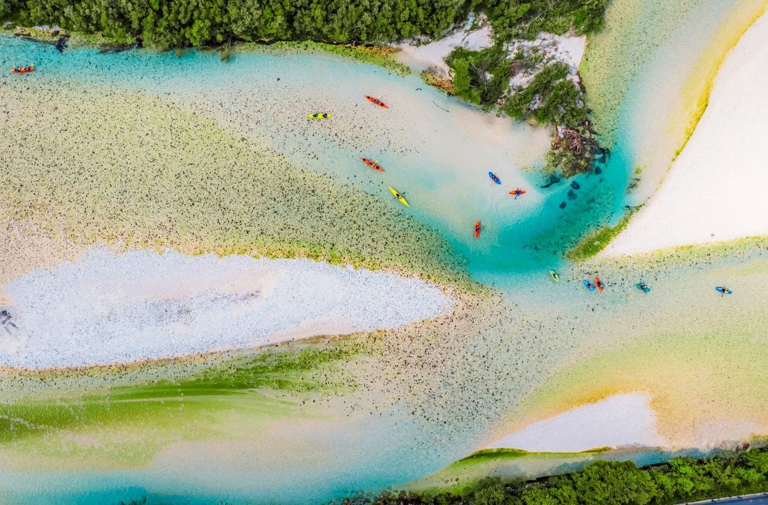 Aerial view of the braided Soča spreading across pale gravel bars with kayakers in the shallow turquoise channels