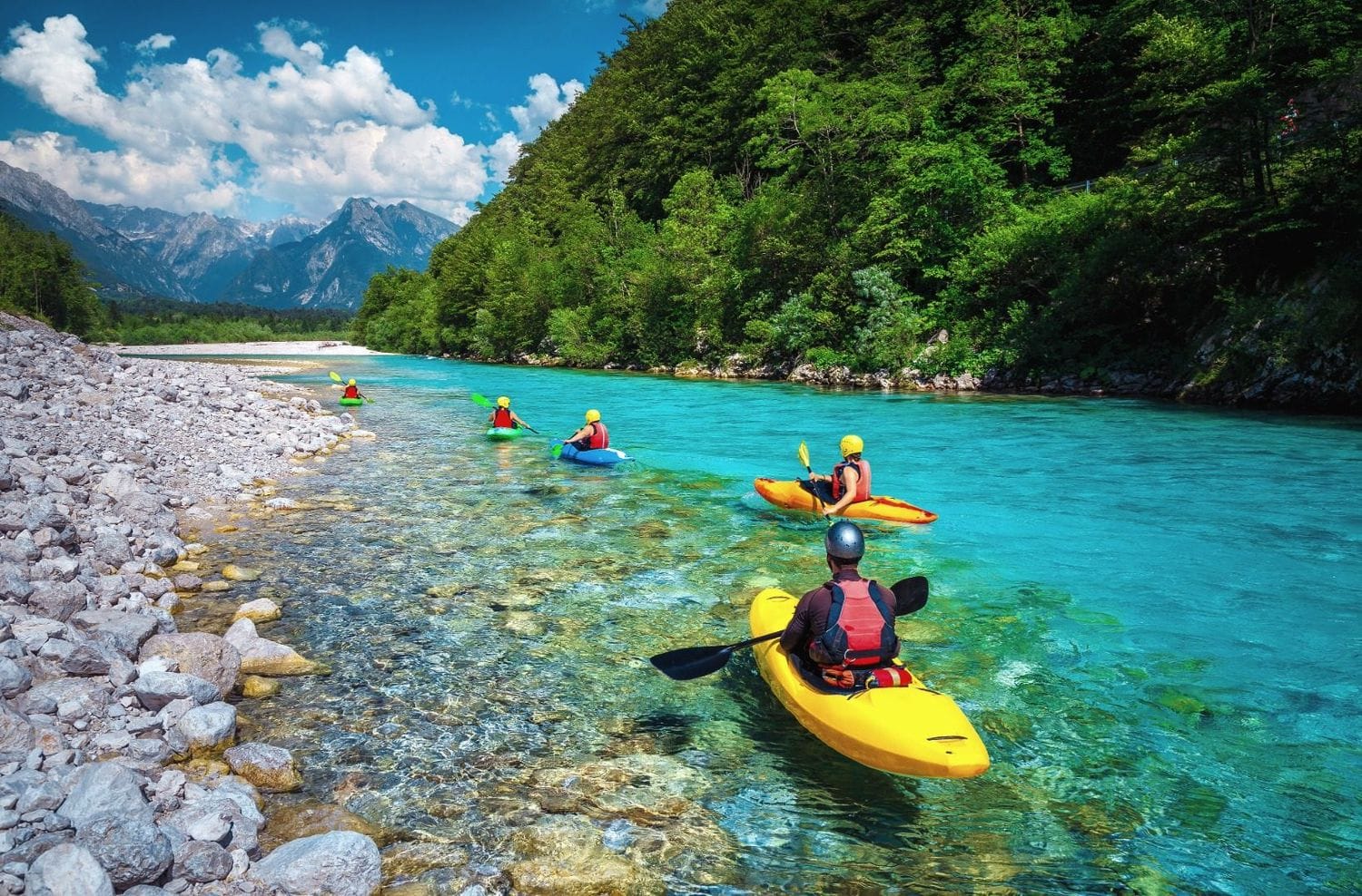 Line of kayakers paddling flat clear turquoise water on the Soča with Julian Alps peaks behind