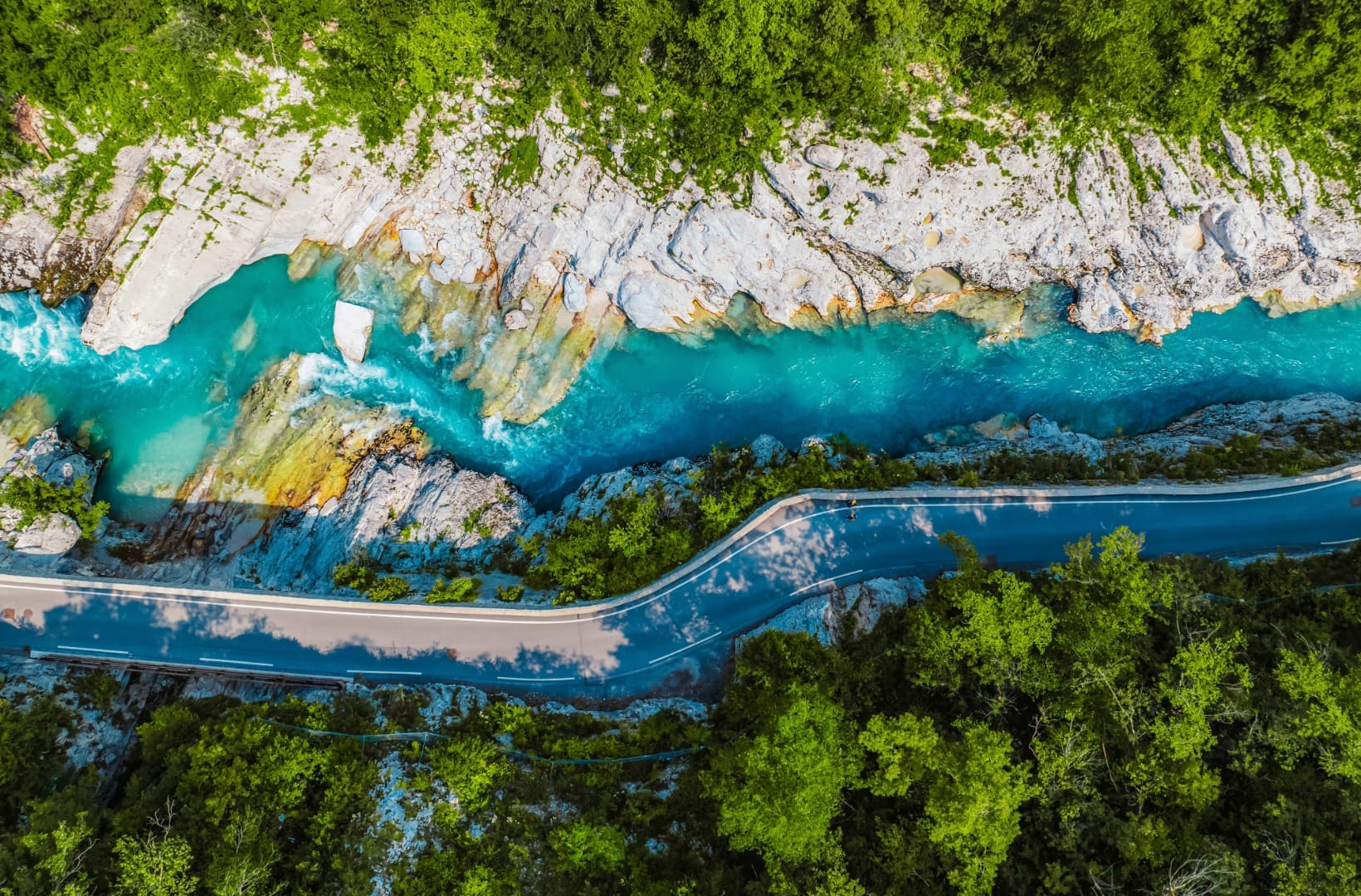 Aerial view of the emerald Soča river running beside a quiet road through steep wooded slopes, a lone cyclist on the tarmac