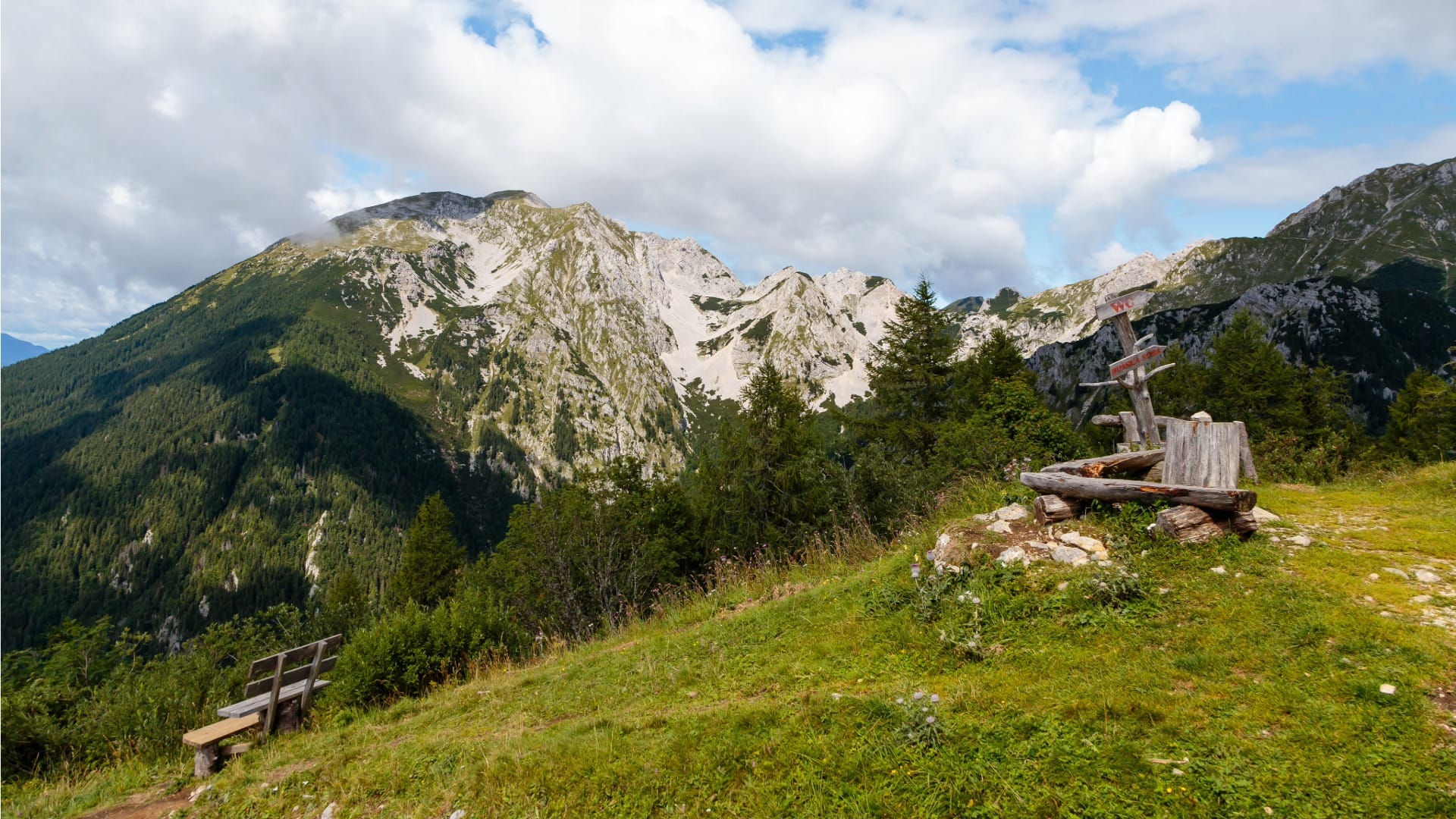 Wooden bench and trail signpost near Prešernova koča na Stolu with the Julian Alps behind