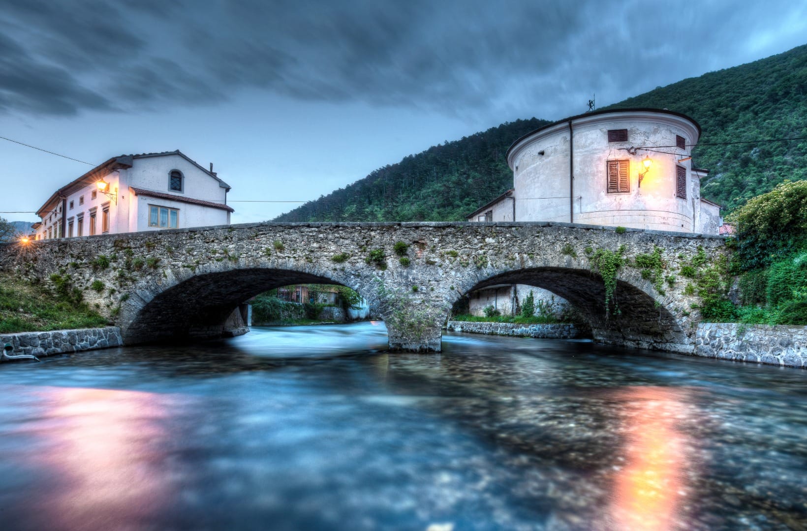 An old stone arched bridge over a branch of the Vipava river in Vipava town, with a round tower building behind