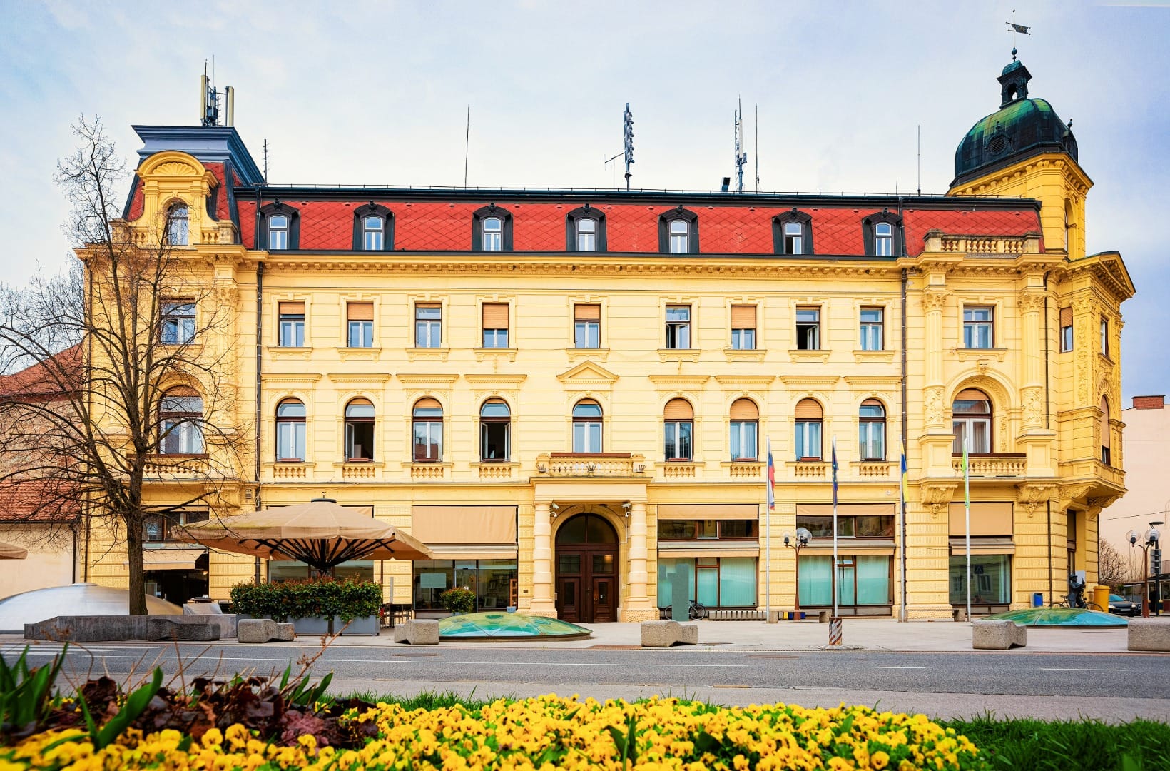 The grand yellow historicist Celjski dom building on a Celje square, with a green copper cupola on its corner tower, a red mansard roof, Slovenian flags and a bed of yellow pansies in front