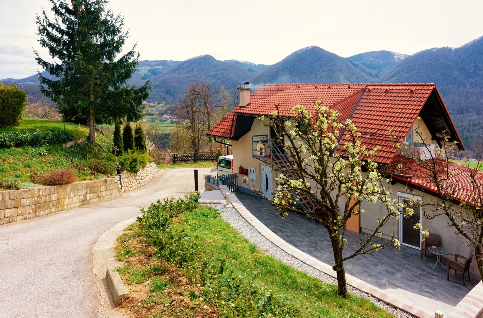 A quiet paved hillside lane curving past a red-roofed house and a low stone wall, a flowering pear tree beside it, looking out over a green valley to rolling forested hills