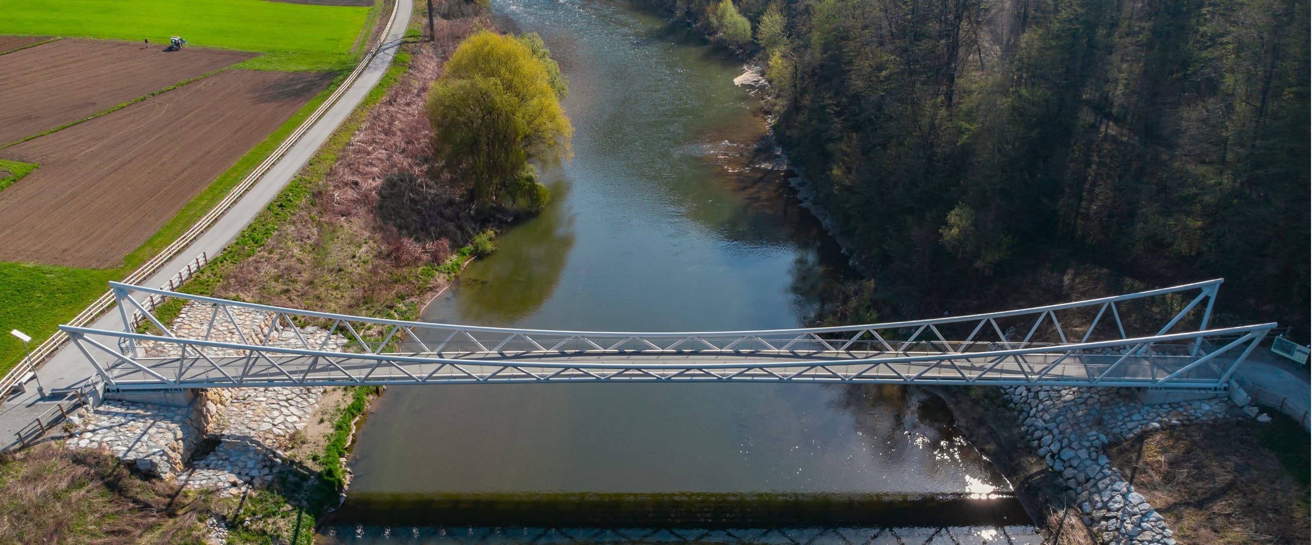 Aerial view of a modern steel-truss cycle and footbridge spanning the Savinja, linking a paved path beside ploughed fields to the wooded far bank