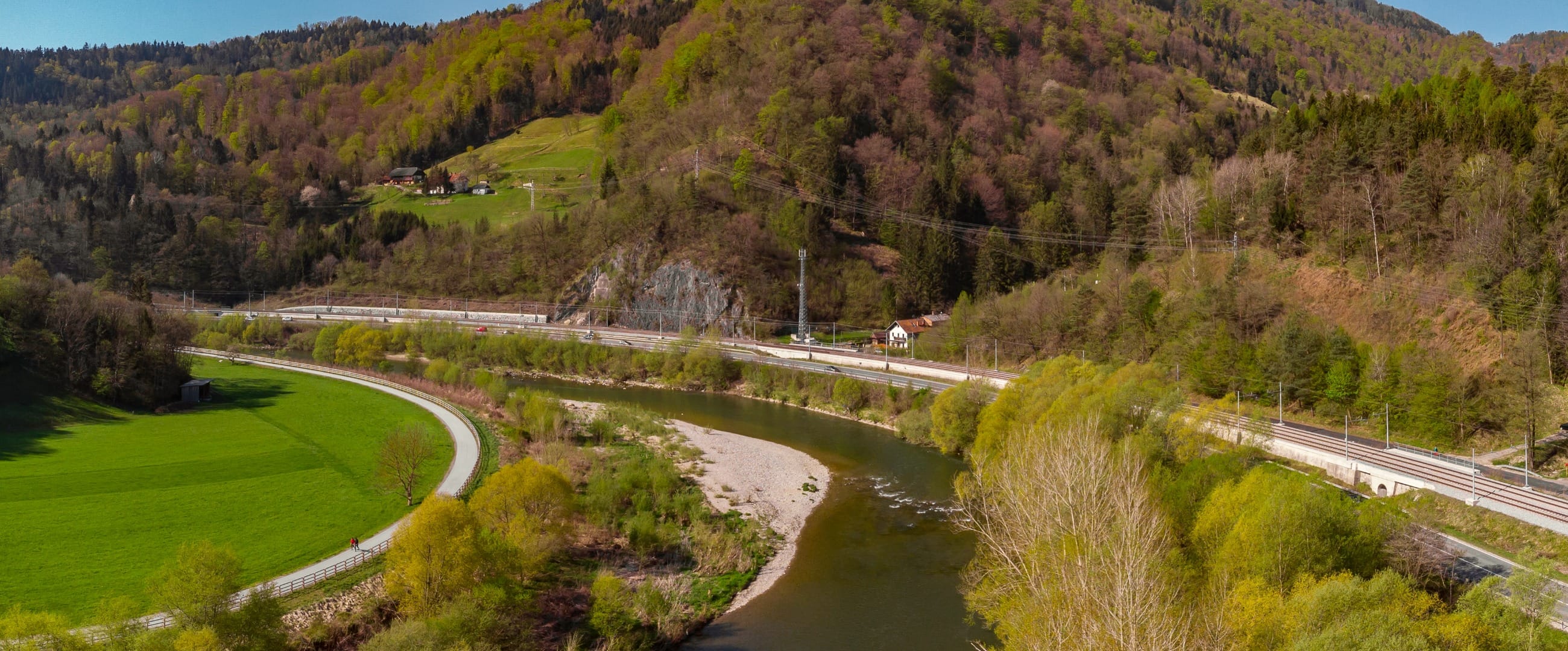 An aerial view of a smooth paved cycle path curving alongside a bright green meadow and the Savinja river, with a road and railway on the far bank and steep wooded hills above