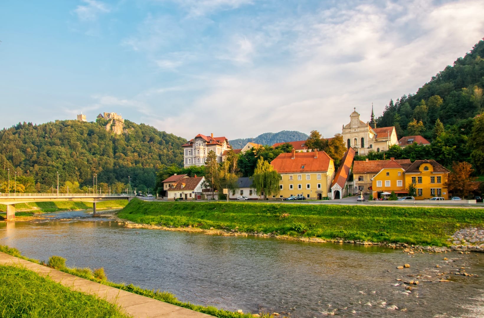 The Savinja river running past pastel townhouses and a church in Celje, with the ruined Old Castle on its wooded hill catching evening light above the town