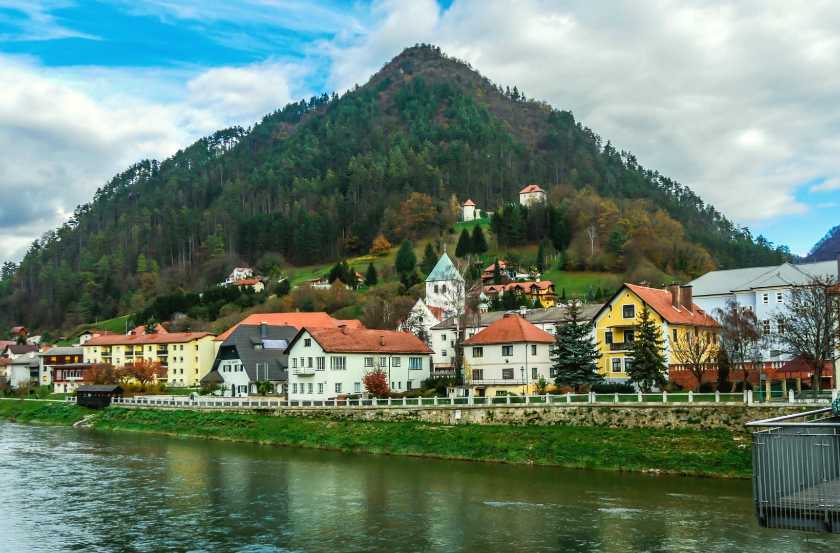 A Savinja-valley town below a steep wooded cone of hill, with a church, pastel riverside houses on a stone embankment and a footbridge over the green river