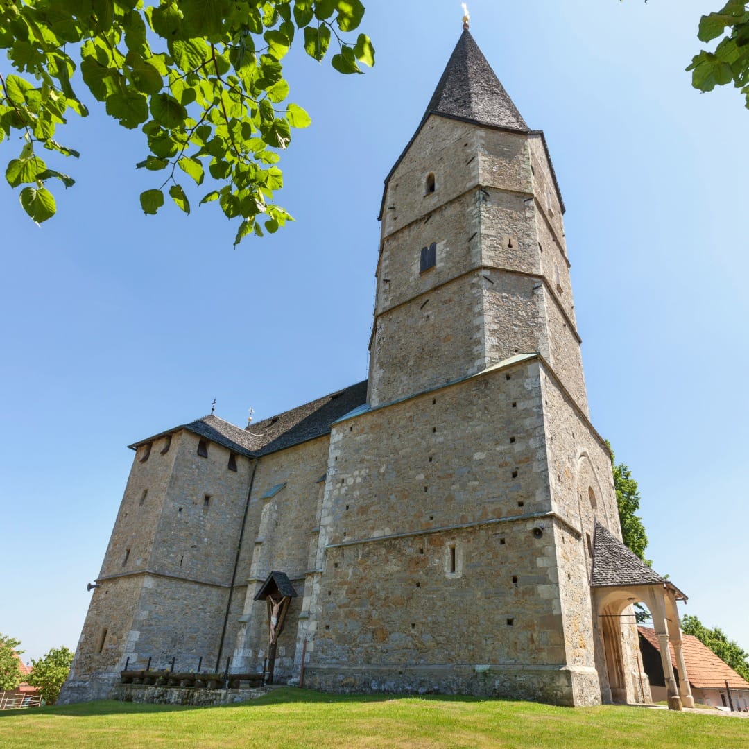 The fortified stone church at Svetina seen from below, a tall octagonal tower with a pyramidal shingled spire, thick buttressed walls and an arched entrance porch, framed by lime branches