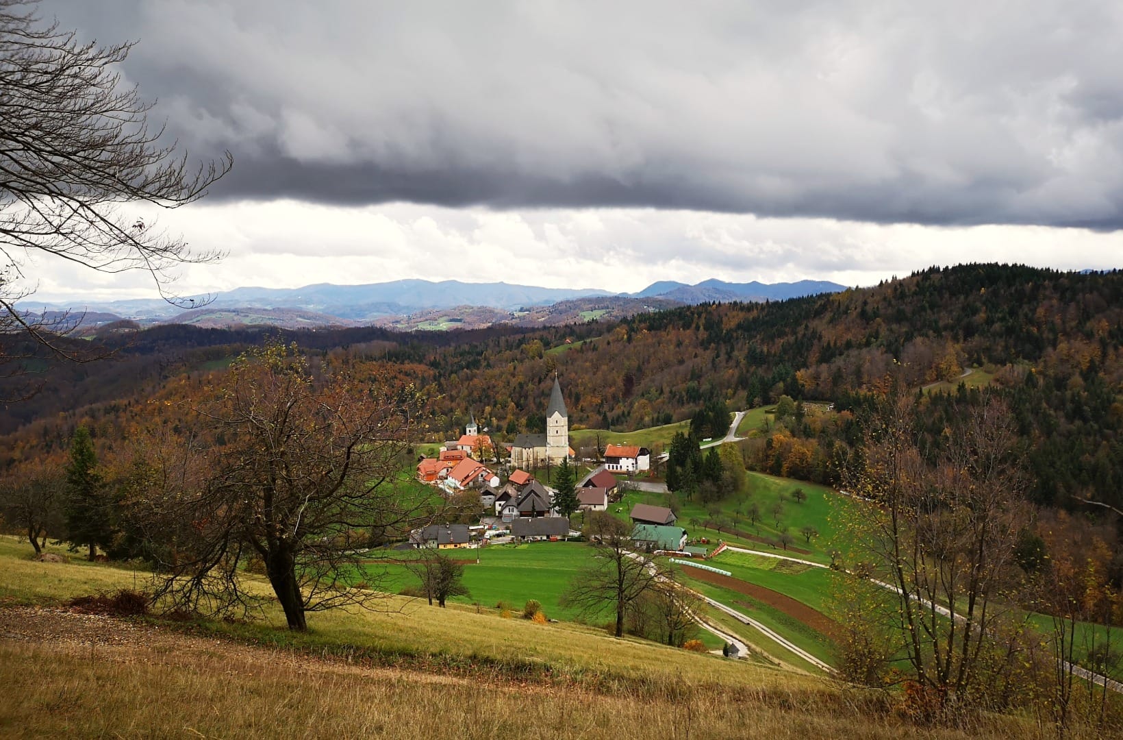 The hilltop hamlet of Svetina with its tall white church tower and a second smaller tower, ringed by houses on a green shoulder, forested hills receding to a mountain horizon under a heavy grey sky