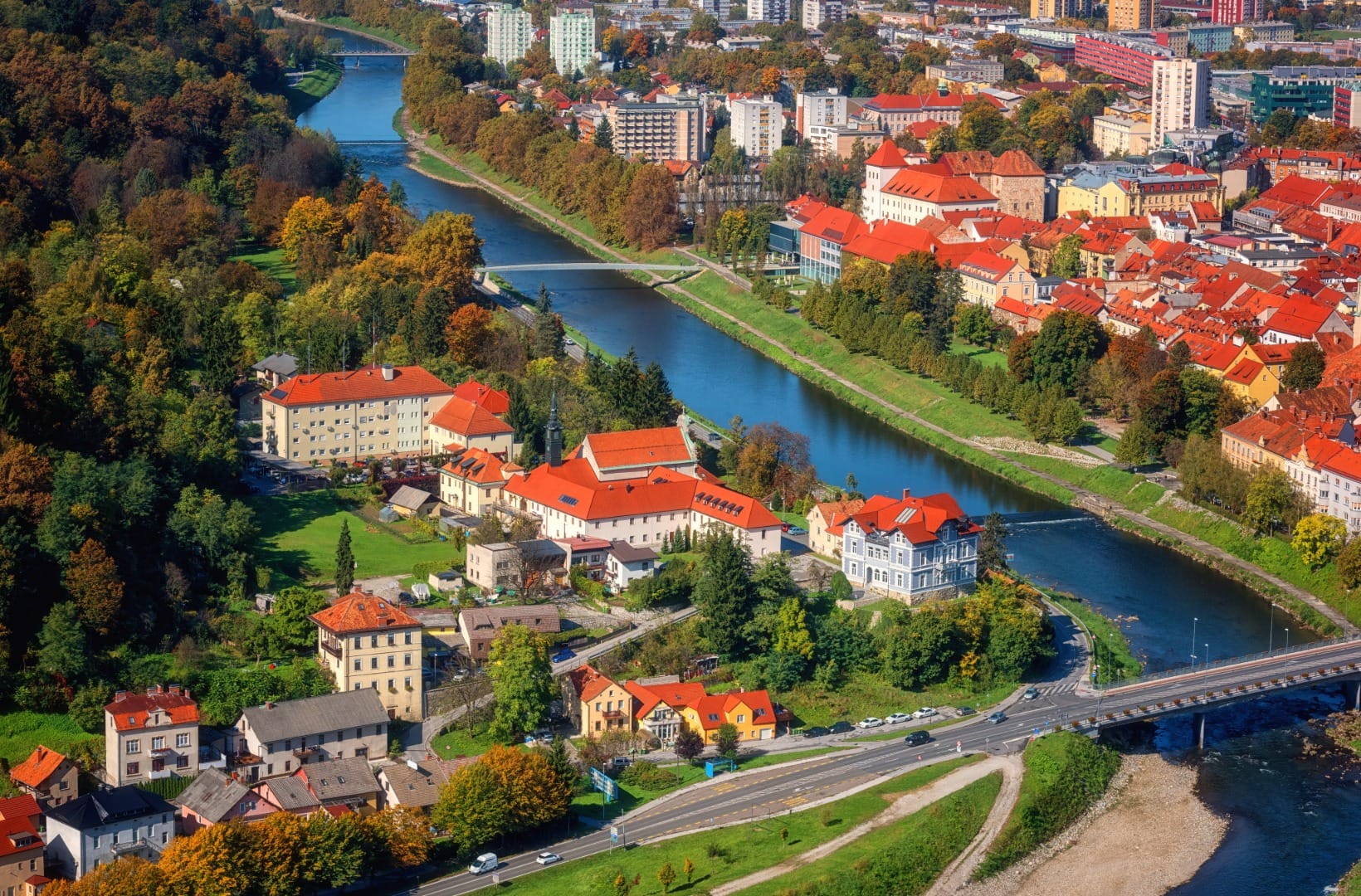 Aerial view over Celje's old town, the Savinja curving through it in a green corridor, red roofs, a pedestrian bridge and apartment blocks behind