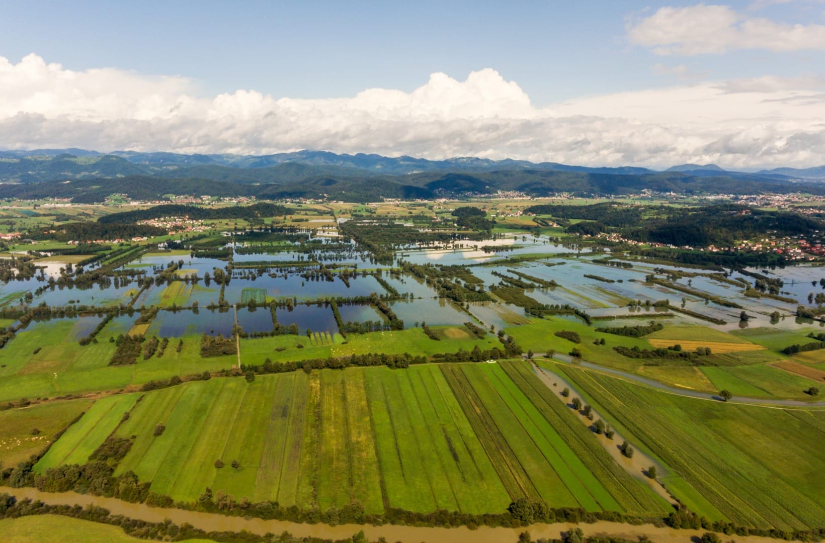 Aerial view of the Ljubljana Marshes with green and flooded fields, a brown river and a railway line, hills on the horizon