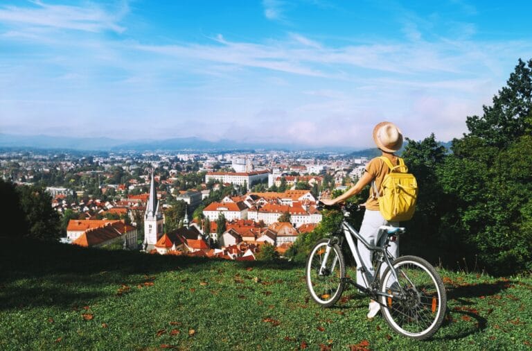 A cyclist standing with a bike on a grassy hill above Ljubljana, looking over the old town rooftops and church spires to the hills beyond