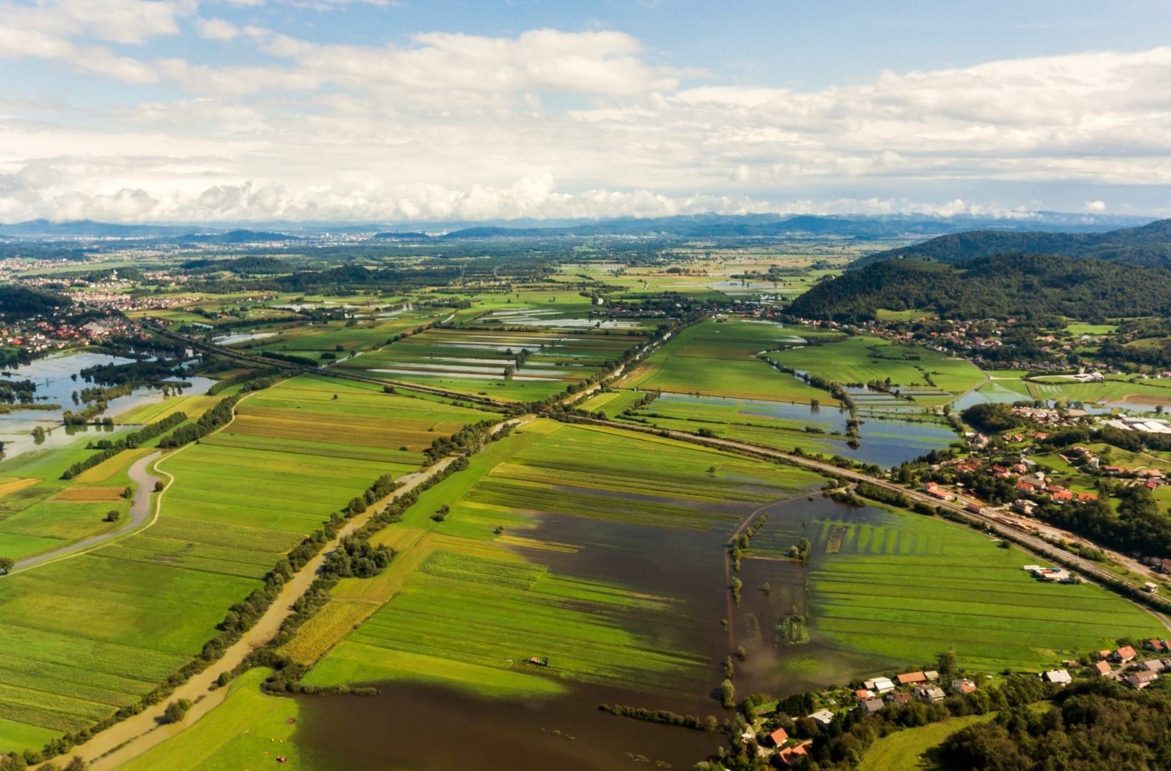 Aerial view of the flat Ljubljana Marshes with flooded hay meadows, hedgerows and a village against low wooded hills