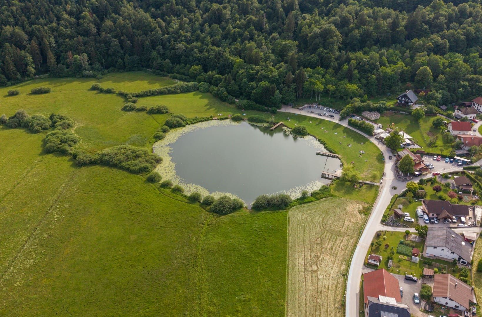 Aerial view of round Podpeč lake with lily pads, a grassy swimming lawn, a lakeside inn and a car park, forest behind
