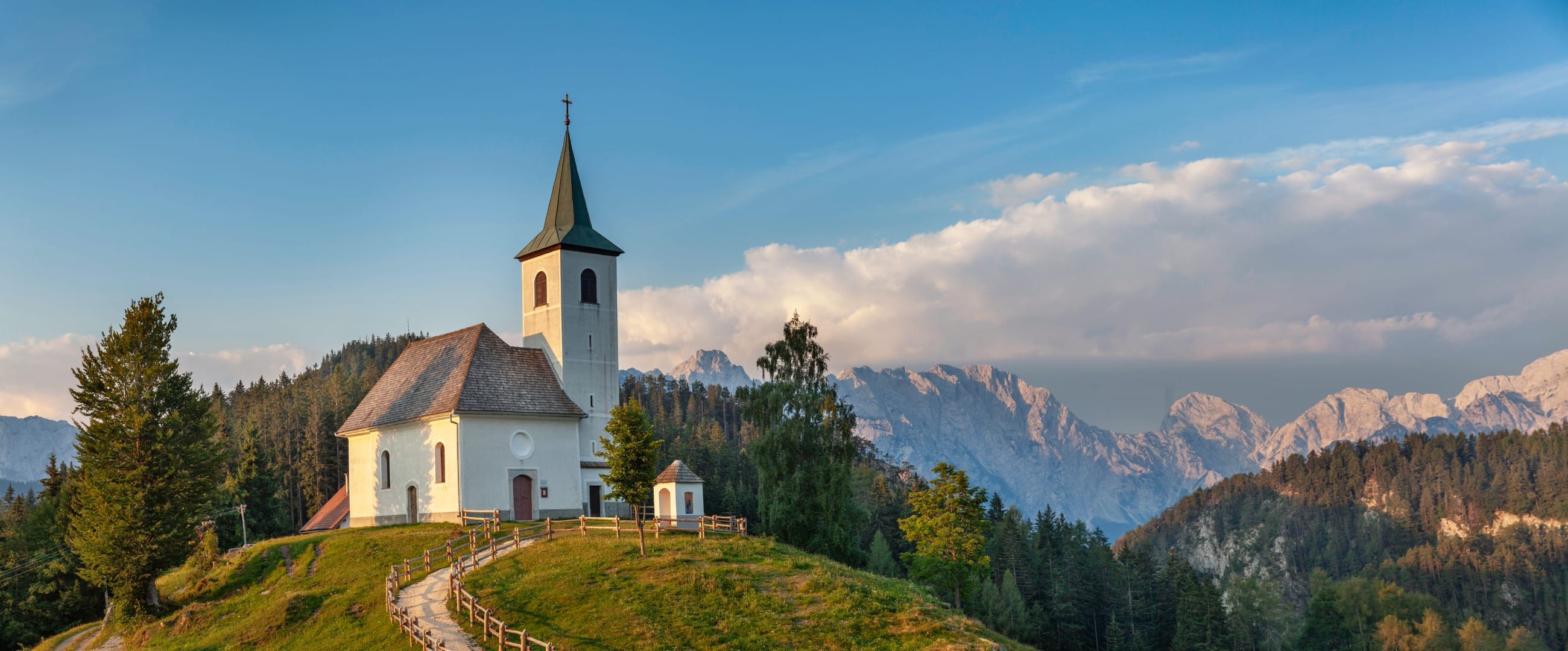The church of the Holy Spirit at Podolševa on its knoll in golden evening light, the Kamnik-Savinja Alps ridge glowing pink behind