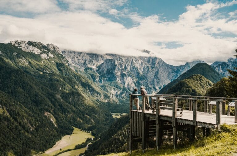 A wooden viewing platform on the Solcava panoramic road with two people looking out over a green valley to the huge grey wall of the Kamnik-Savinja Alps