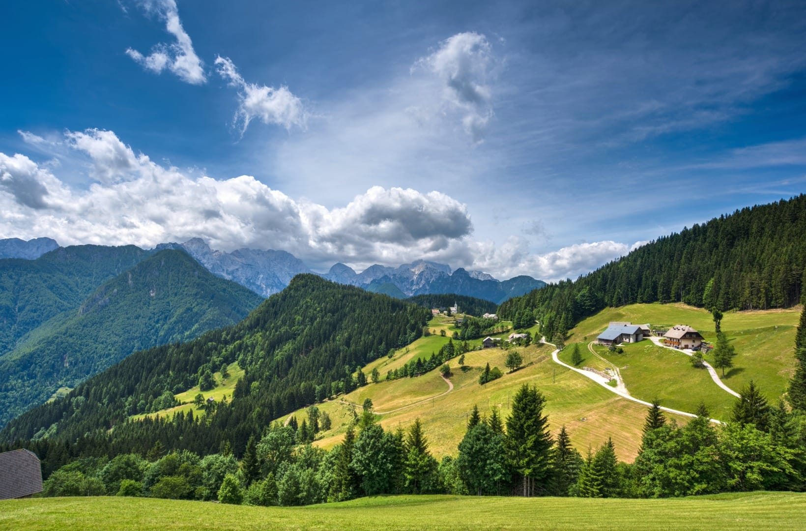 Rolling green pastures with scattered white farmhouses and a small church, forested ridges and the grey Alps wall in the distance
