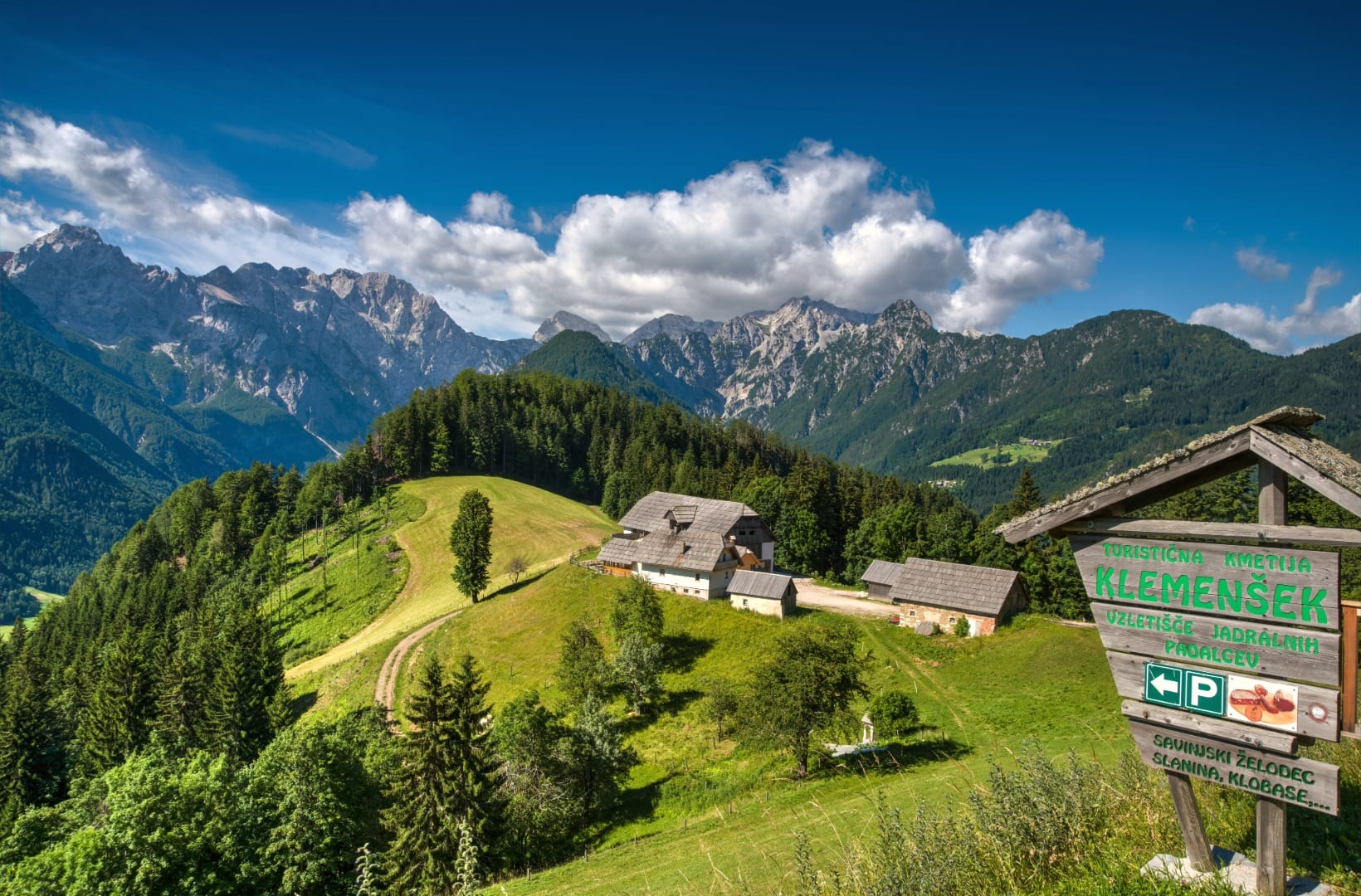 The Klemenšek mountain farm on a grassy shelf with its wooden sign advertising cured meats, the jagged Kamnik-Savinja Alps ridge behind