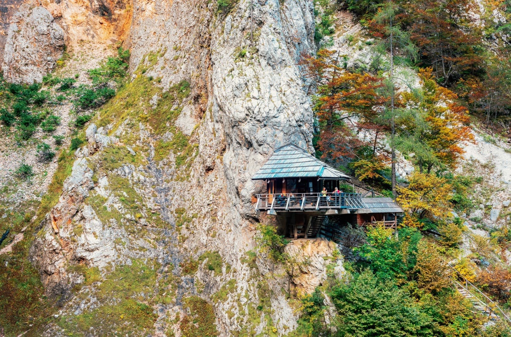 Close view of the Orlovo gnezdo hut clinging to the cliff face among autumn larches, people on its terrace