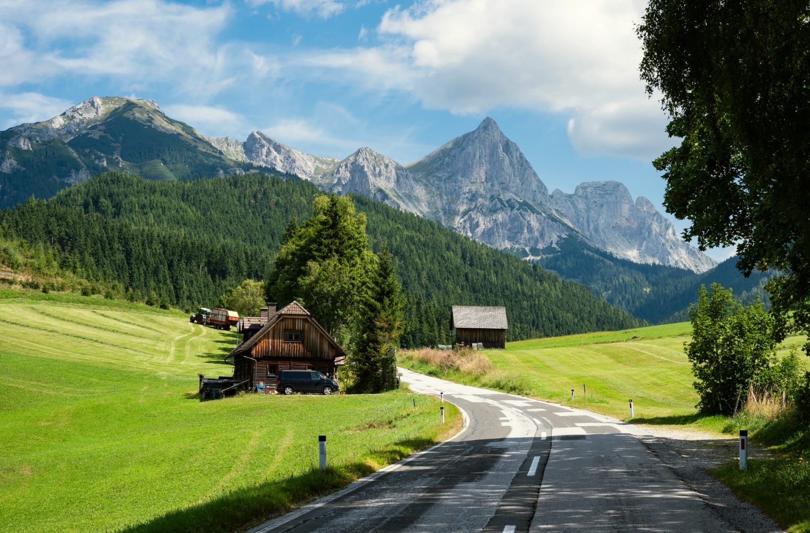 A quiet paved road curving past a wooden mountain farmhouse and hay barn in green meadows below the sharp grey peaks of the Kamnik-Savinja Alps