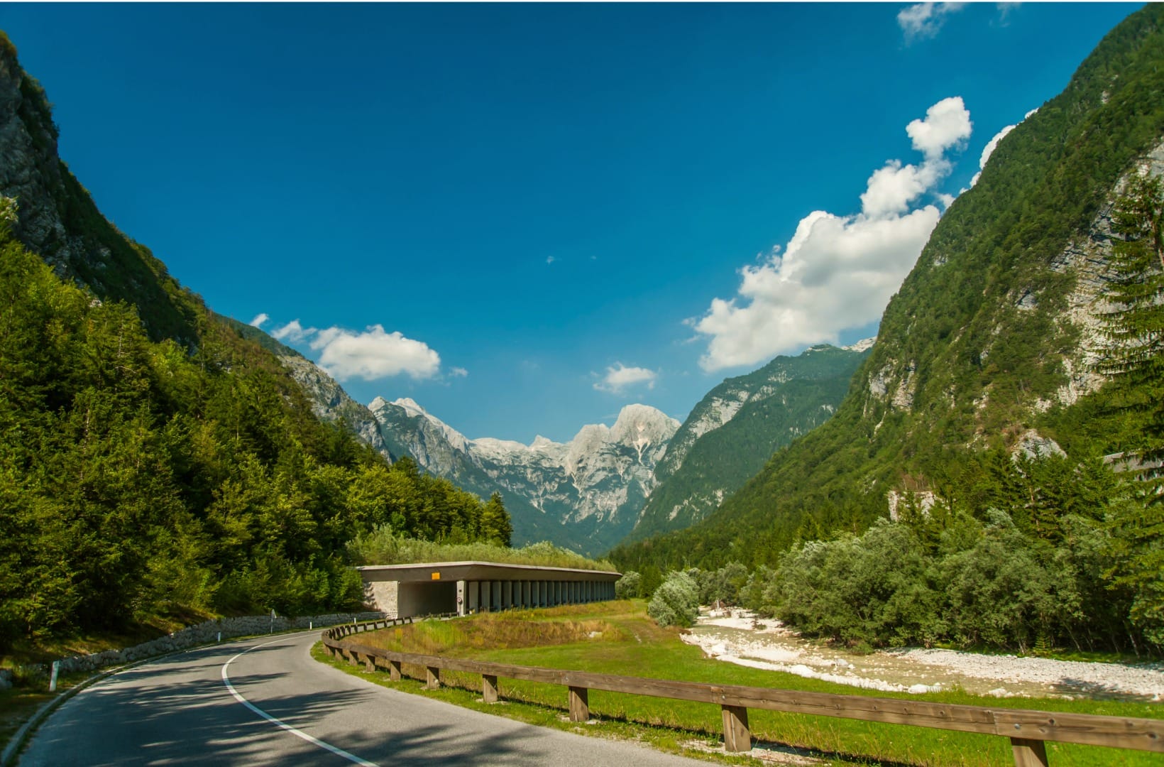 Regional road 206 passing a concrete open-sided avalanche gallery in the Soča valley with white limestone peaks under a blue sky