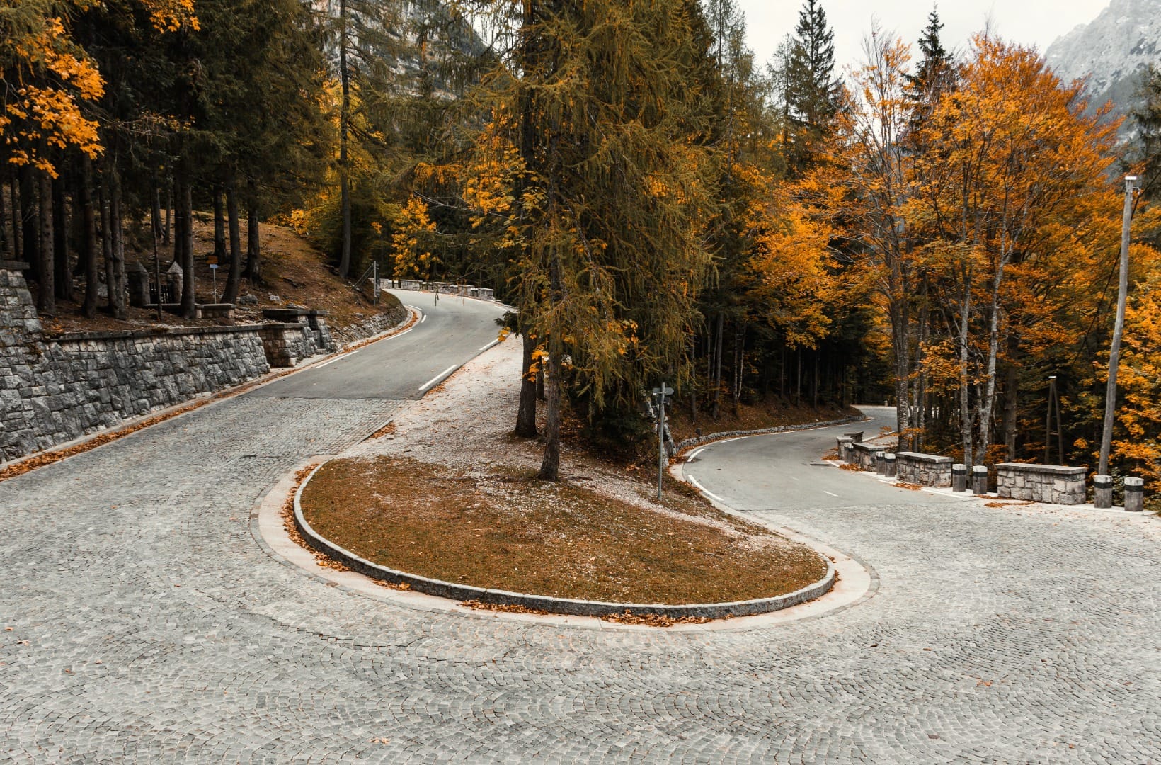 A wide cobbled hairpin bend on the north side of Vršič with a grassy island and a larch in the middle, autumn colour, stone retaining wall