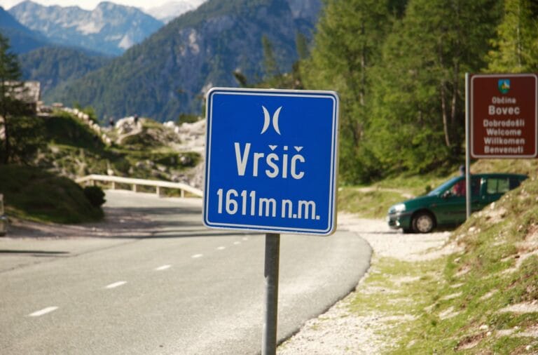 A blue road sign reading "Vrsic 1611 m n.m." at the Vrsic pass, mountains behind and an Obcina Bovec welcome sign
