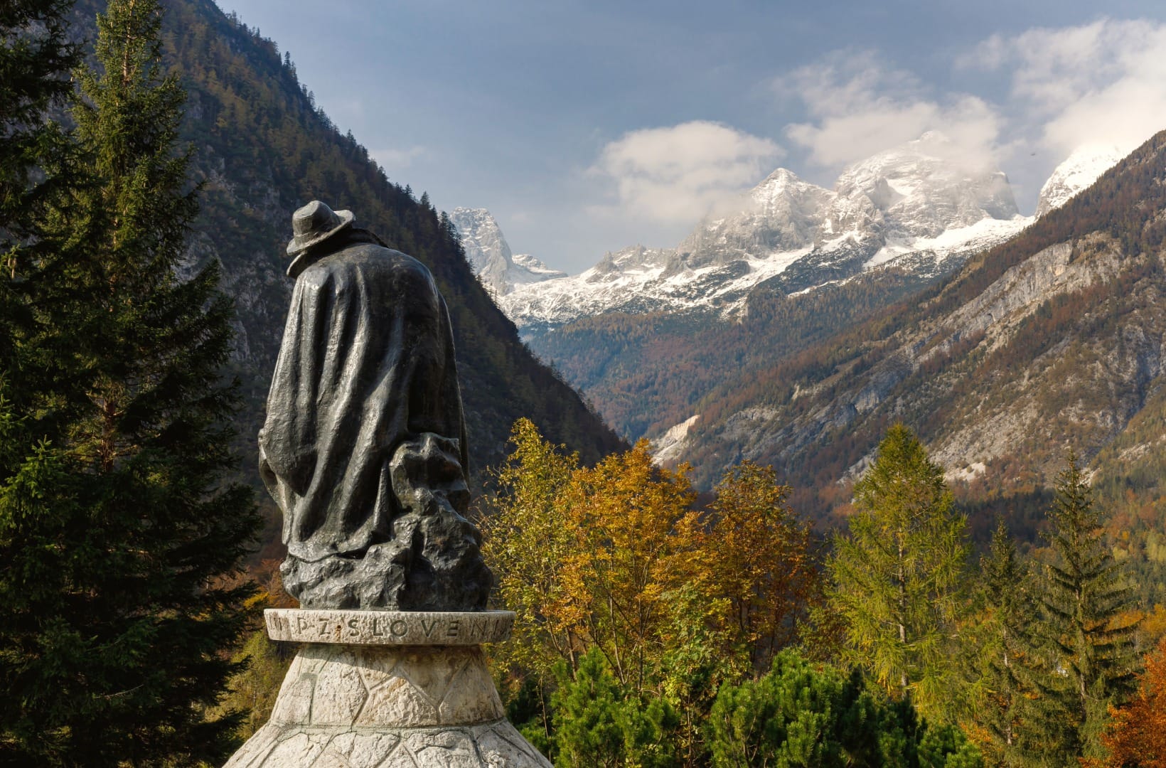 The bronze monument to Julius Kugy, a caped, hatted figure seen from behind, looking down the autumn Trenta valley toward snow-dusted peaks