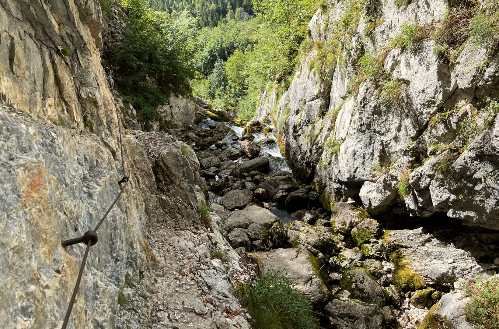 A narrow limestone gorge on the upper Soča with a fast stream over pale boulders and a steel guide cable bolted into the rock wall