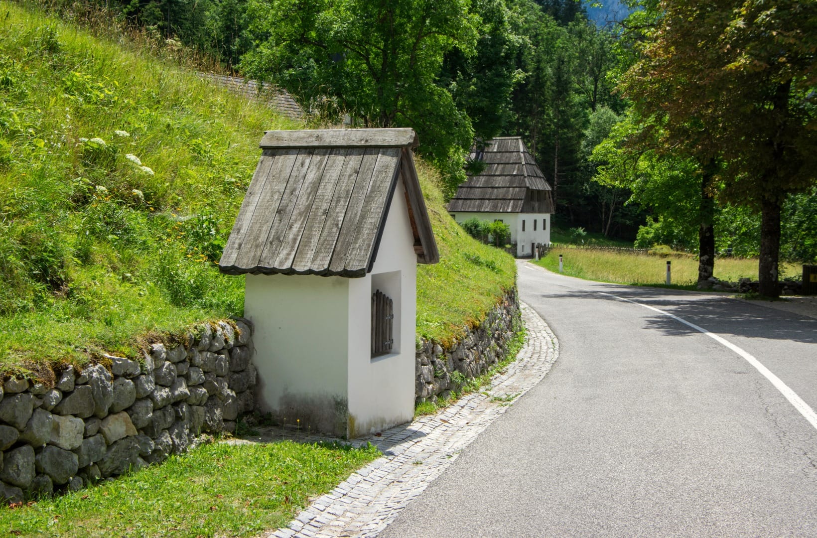 A small white wayside shrine with a wood-shingle roof beside the road in Trenta, shingle-roofed houses behind
