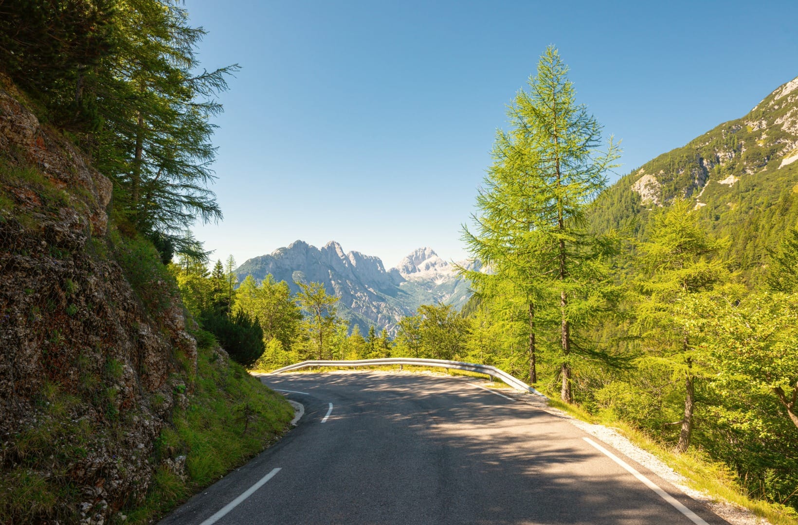A sweeping paved bend on the upper Vršič climb with larch trees and jagged limestone peaks framed ahead