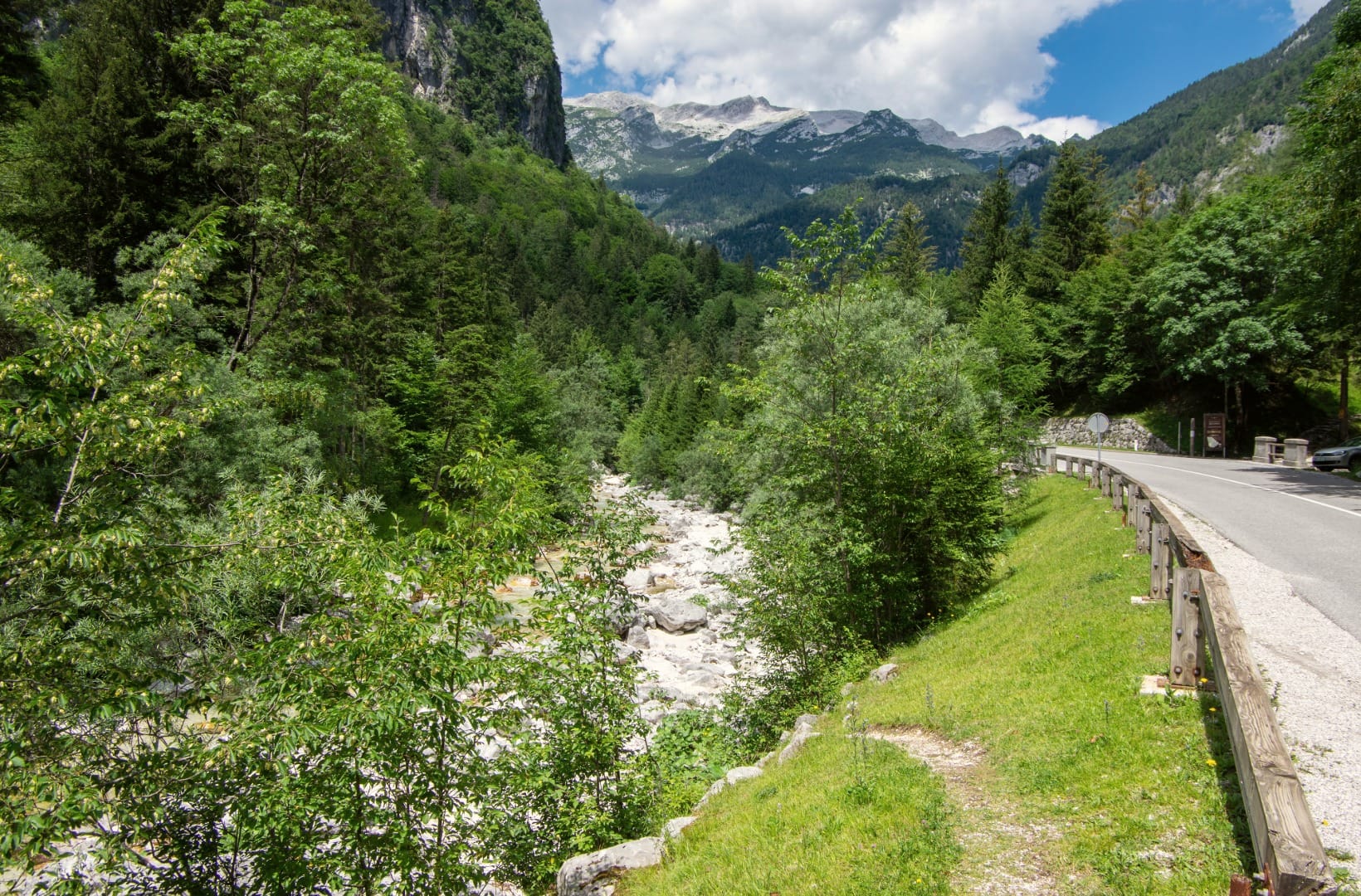 Regional road 206 curving through the wooded Soča valley with the young river in its white-boulder bed and snow-streaked peaks ahead